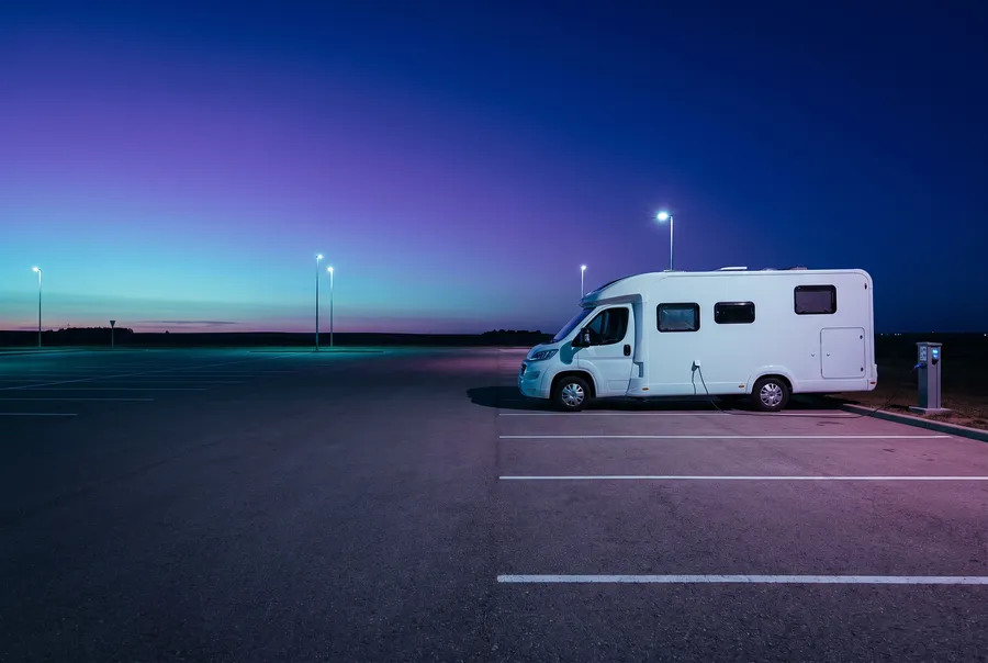 Generated picture: a white motorhome parked at dusk on a wide empty lot, its power cord running to a grey electrical pedestal, prairie sky behind.